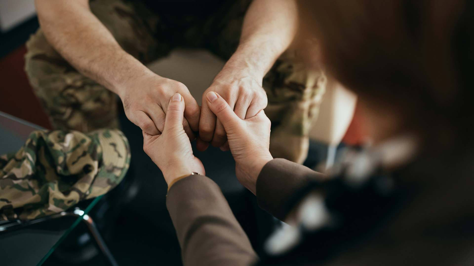 Two pairs of hands holding each other, with a person in military uniform in the background.